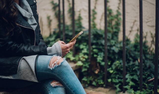 figura femminile con jeans strappati e giubbino di pelle che tiene uno smartphone in mano