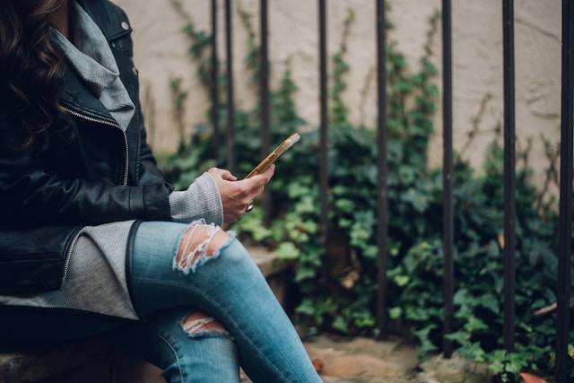 donna con jeans strappati e giubbino di pelle che tiene uno smartphone in mano.